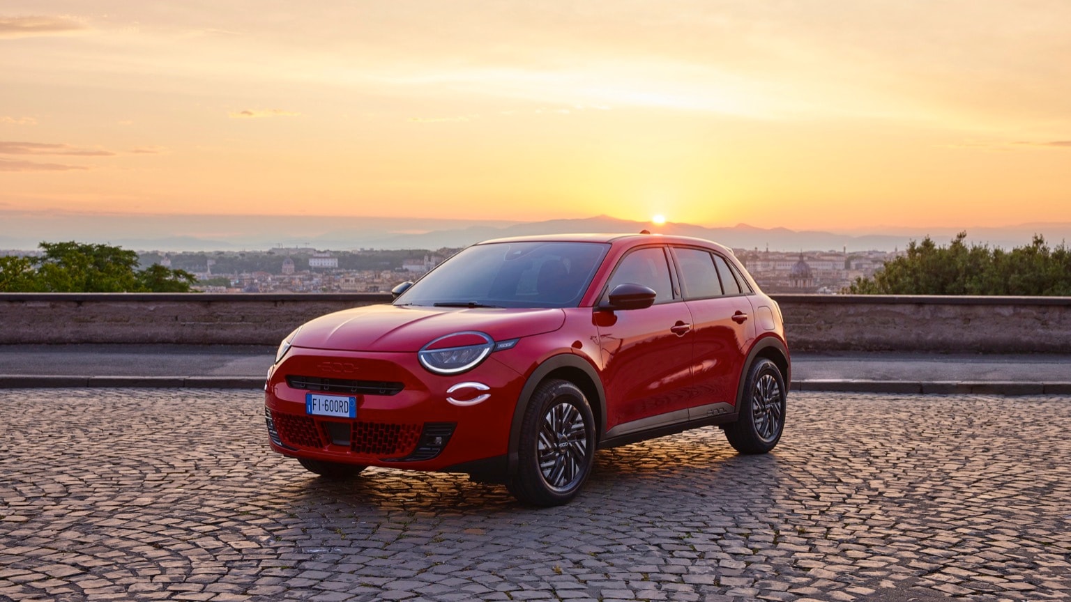 une voiture rouge garée sur une route en briques avec un coucher de soleil en arrière-plan
