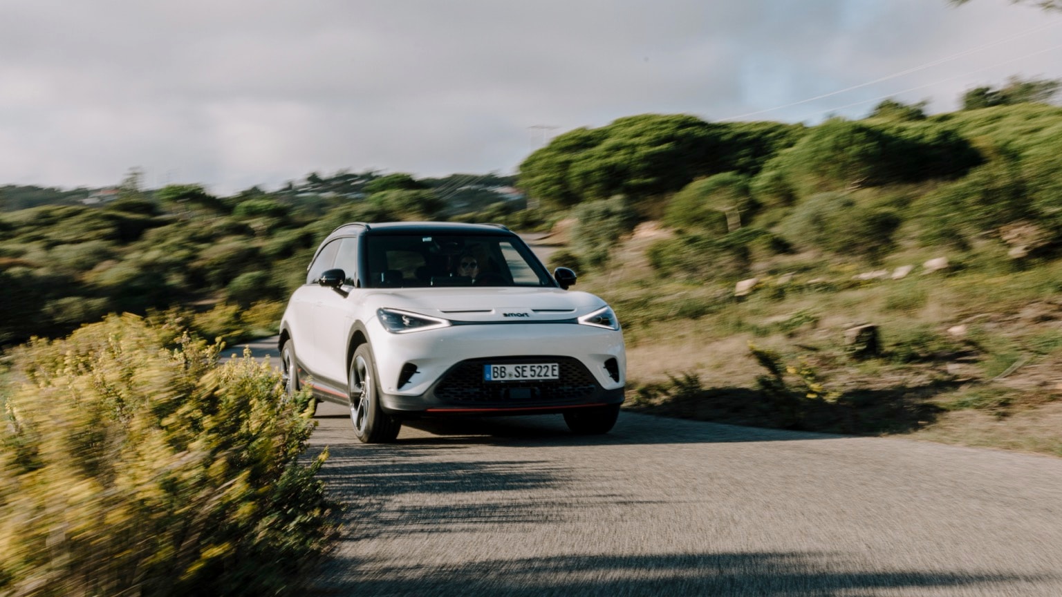 a white car driving on a road with bushes and trees in the background
